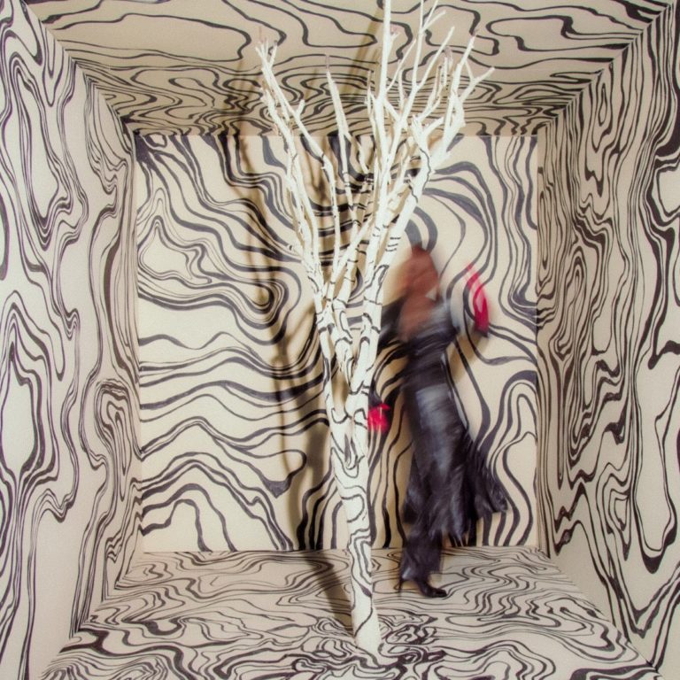 Unscharfe Frau in schwarzem Kleid und roten Handschuhen tanzt in einem surrealen Raum mit psychedelisch gemusterter Tapete und einem weißen, kahlen Baum. Alles wirkt wie Teil einer optischen Illusion.