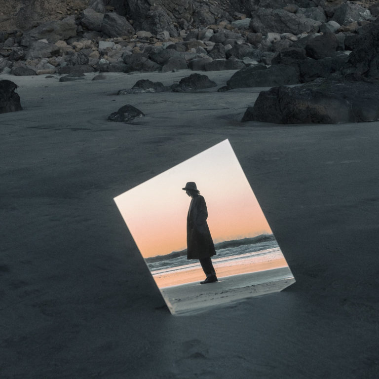 Ein Spiegel lehnt leicht geneigt im dunklen Sand einer felsigen Küstenlandschaft. Darin spiegelt sich eine Person im Mantel bei Sonnenuntergang am Meer.