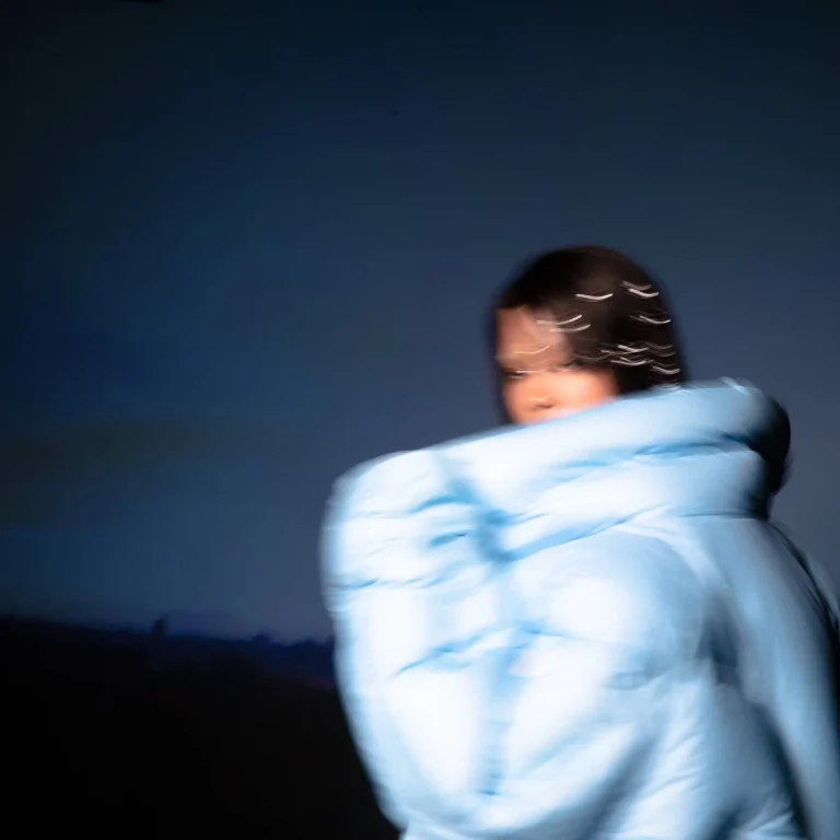 Verschwommenes Porträt von Shygirl in einer übergroßen, hellblauen Jacke vor dunklem Hintergrund – das Cover ihres vielschichtigen Debütalbums Nymph.