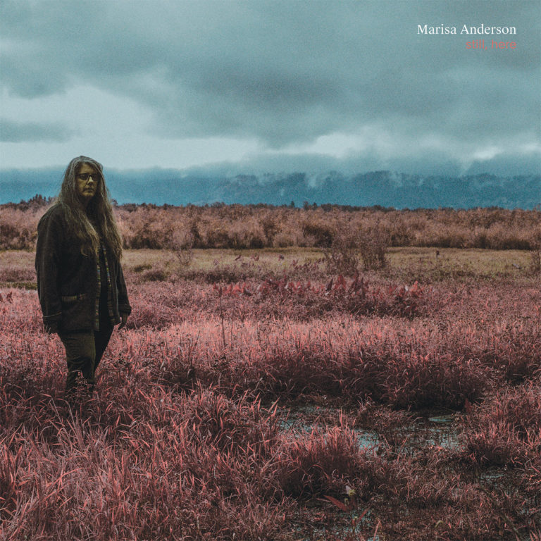 Marisa Anderson steht allein in einer kargen, rötlich verfärbten Landschaft unter schwerem Himmel – das Cover ihres introspektiven Gitarrenalbums Still, Here.