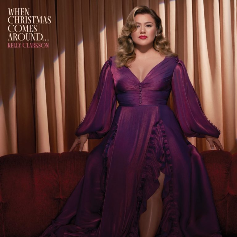 Albumcover von Kelly Clarkson – „When Christmas Comes Around...“: Die Sängerin sitzt im violetten Glamourkleid auf einem roten Sofa vor goldfarbenem Vorhang.
