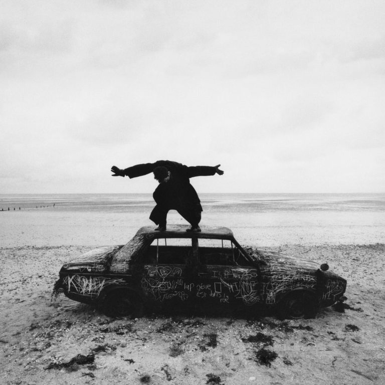 Schwarz-weiß-Foto einer Person, die auf einem alten, bemalten Autowrack am Strand balanciert – ein ausdrucksstarkes Bild zwischen Verfall und Freiheit.