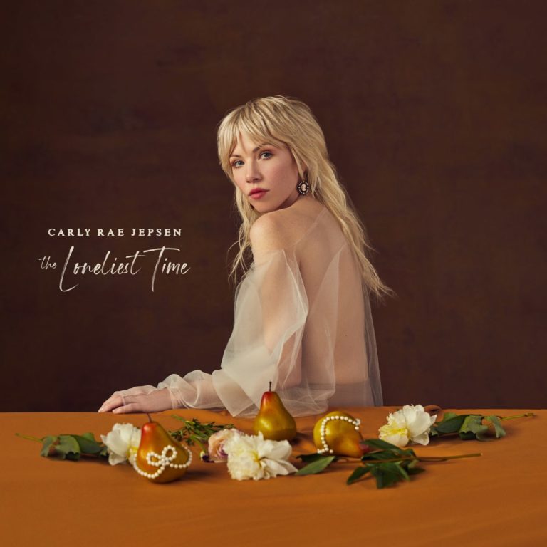 Carly Rae Jepsen sitzt in einem eleganten, halbtransparenten Kleid an einem gedeckten Tisch mit Früchten und Blumen, blickt verträumt und nachdenklich über ihre Schulter – Cover ihres Albums „The Loneliest Time“.