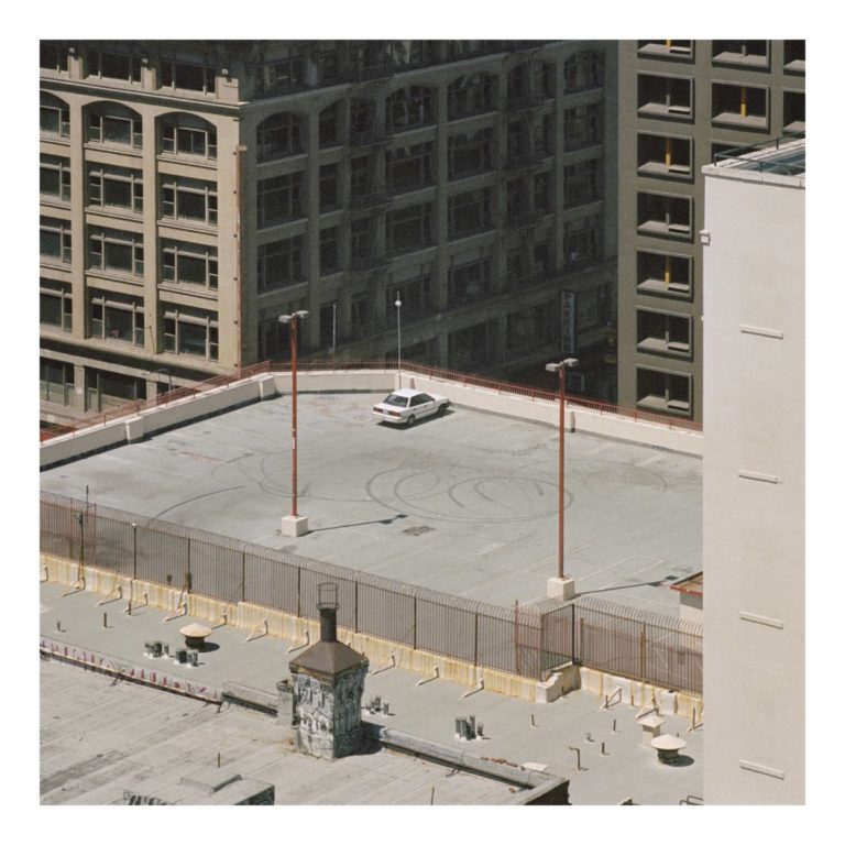 Ein einsames Auto steht verlassen auf dem Dach eines städtischen Parkhauses, umgeben von großen Gebäuden – Cover des Albums „The Car“ von Arctic Monkeys.
