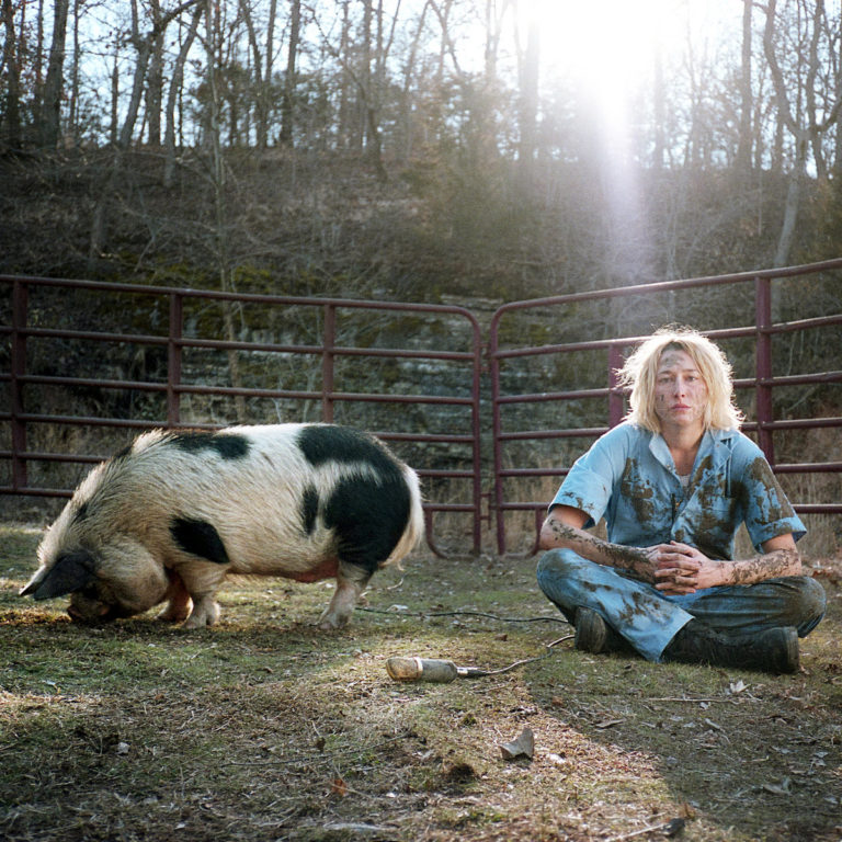 Eine verschmutzte Musikerin sitzt neben einem suhlenden Schwein im staubigen Gehege, im Rücken rostige Zäune und kahles Waldstück, die Sonne blitzt durch.