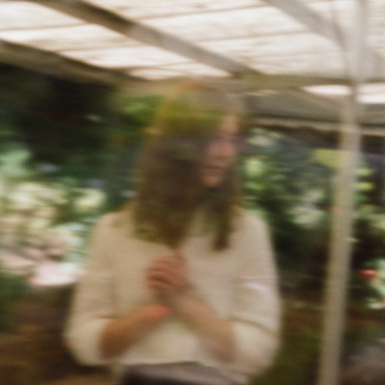 Verschwommenes Porträt einer Frau hinter Glas, in gedecktem Licht mit natürlichem Hintergrund – träumerisch, introvertiert, atmosphärisch.