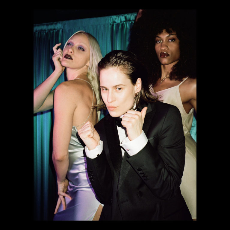 Christine and the Queens im Smoking mit zwei glamourösen Tänzerinnen im Satin-Kleid vor türkisgrünem Vorhang – stilisiert, queer, performativ.