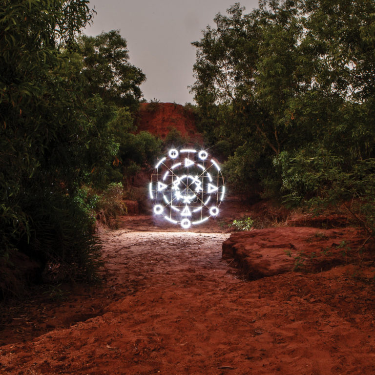 Ein hell leuchtendes, geometrisches Symbol mit ethnisch-ritueller Ästhetik schwebt inmitten einer rötlichen, trockenen Schluchtlandschaft, umgeben von dichtem Grün. Die Szenerie wirkt zugleich außerweltlich und verwurzelt – wie ein geheimnisvolles Portal in Raum und Zeit.