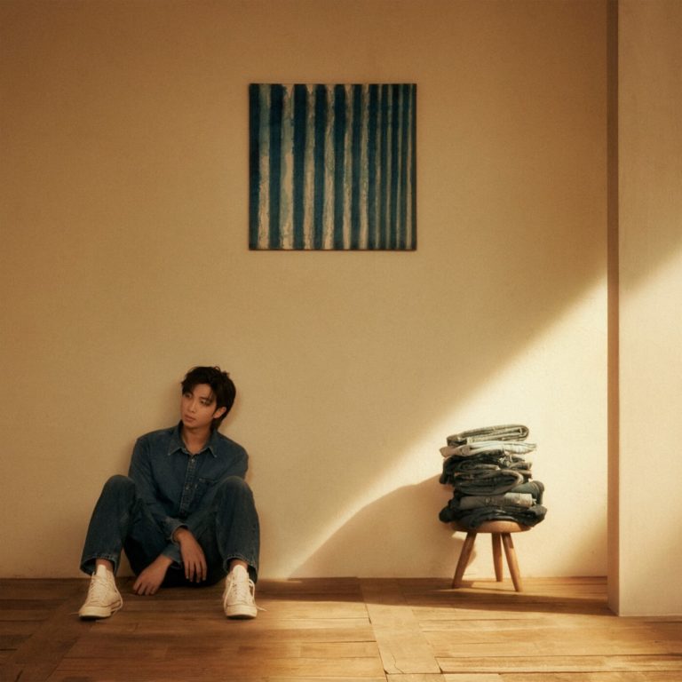 RM sitzt in einem minimalistischen Raum aus Holz und warmem Licht, Denim auf Denim, den Blick ins Leere gerichtet – ein ruhiges, nachdenkliches Setting unter einem abstrakten Gemälde, das sein Soloalbum Indigo visuell einleitet.