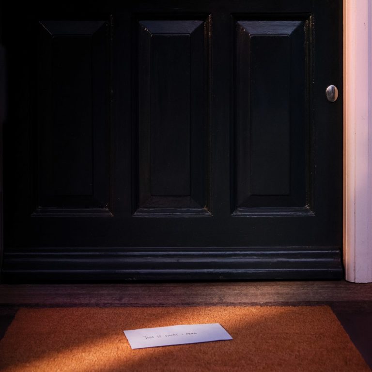 Schwarze Haustür bei Dämmerlicht, davor ein Umschlag auf der Fußmatte mit der Aufschrift „This Is What I Mean“ – ein schlichtes, symbolstarkes Albumcover, das Intimität und Ankunft andeutet.