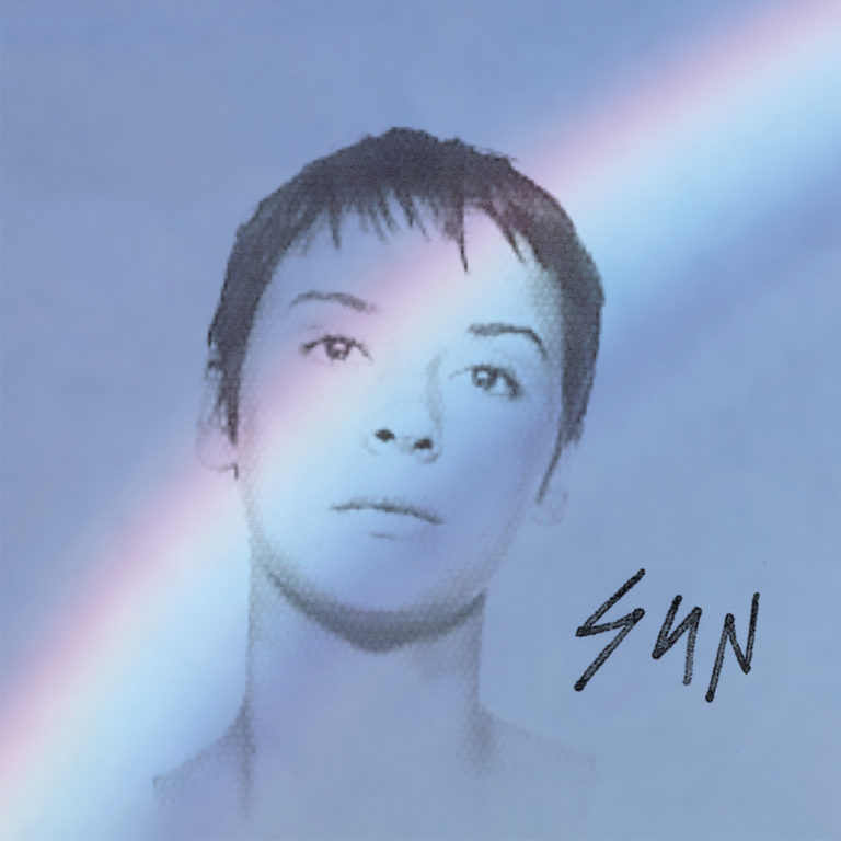 Porträt einer Frau mit kurzem Haar und Regenbogenlicht auf ihrem Gesicht vor blauem Hintergrund.