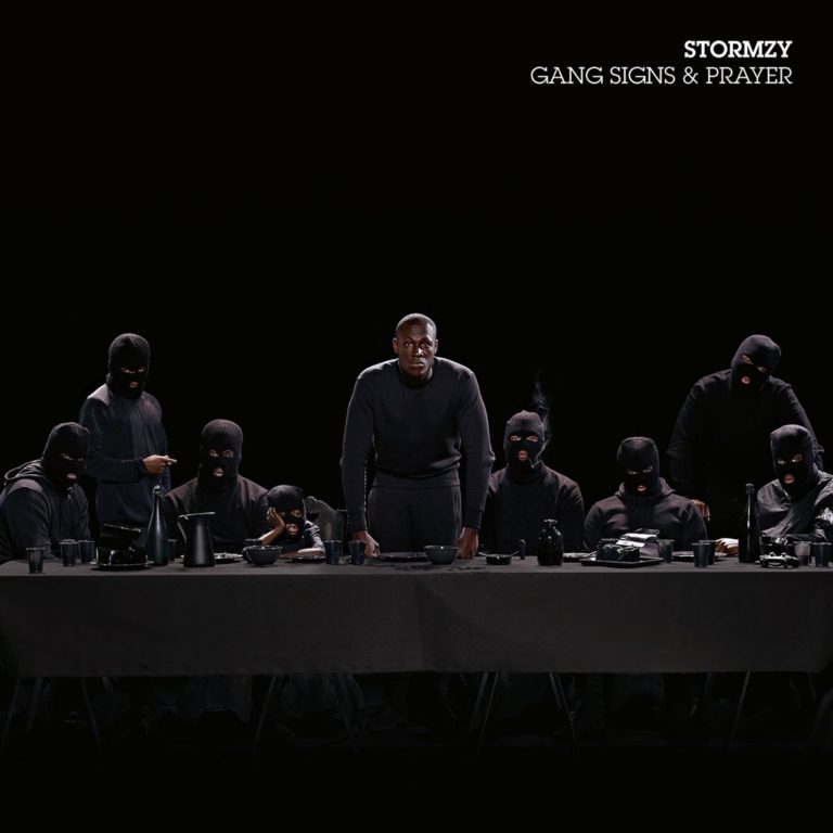 Stormzy steht in schwarzer Kleidung an einem Tisch, umgeben von maskierten Personen.