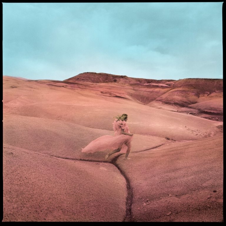 Albumcover von „Strays“ mit Margo Price in rosafarbenem Kleid, allein in einer surrealen, rosa eingefärbten Wüstenlandschaft unter pastellblauem Himmel – ätherisch und poetisch.