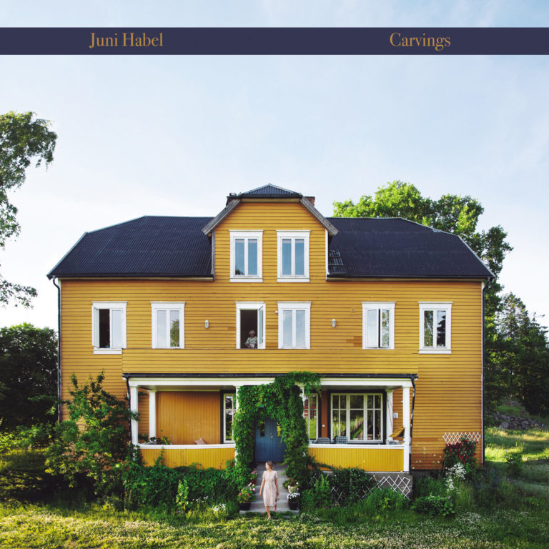 Albumcover von „Carvings“ von Juni Habel mit einem großen gelben Holzhaus, vor dem eine Frau im Sommerkleid steht – ländlich, poetisch und melancholisch.