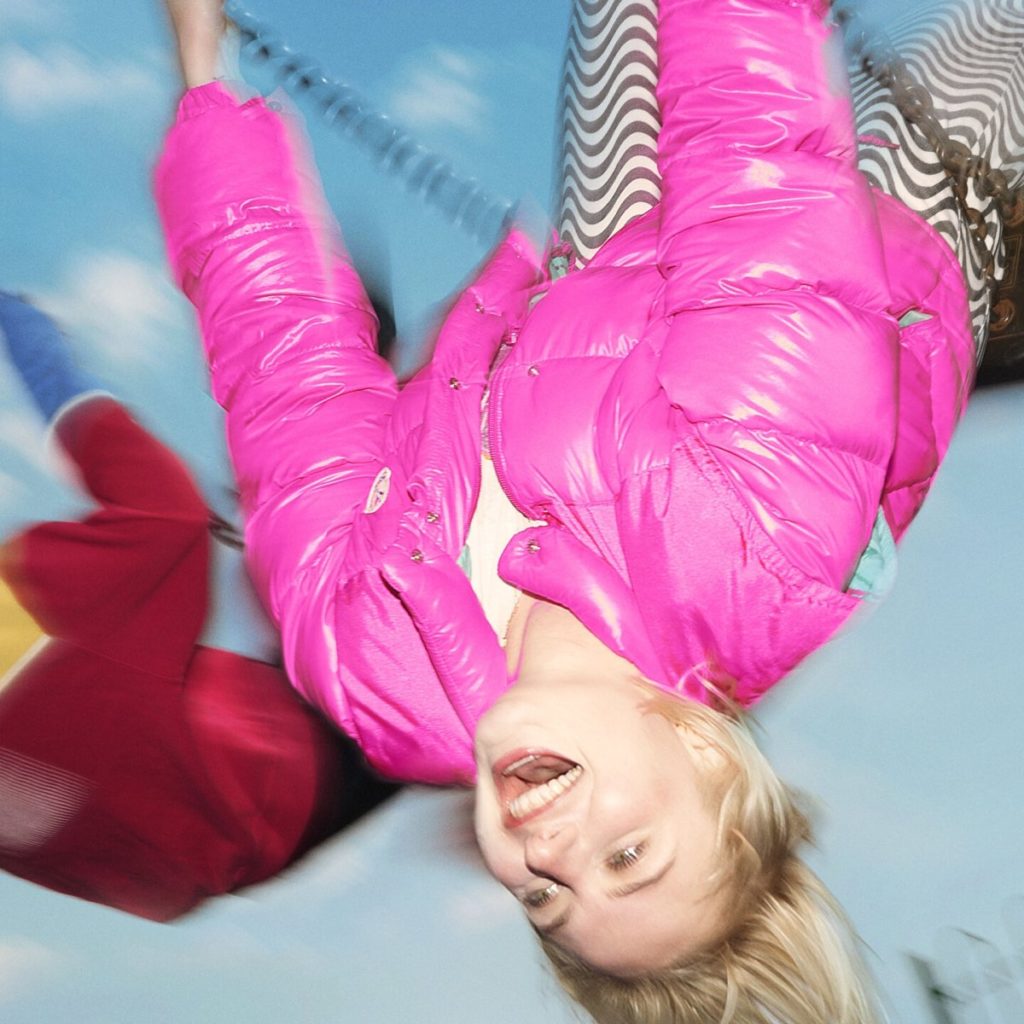 Låpsley lachend in einem Karussell, in pinker Jacke kopfüber fotografiert – Cover von „Cautionary Tales of Youth“, dynamisch und verspielt inszeniert.