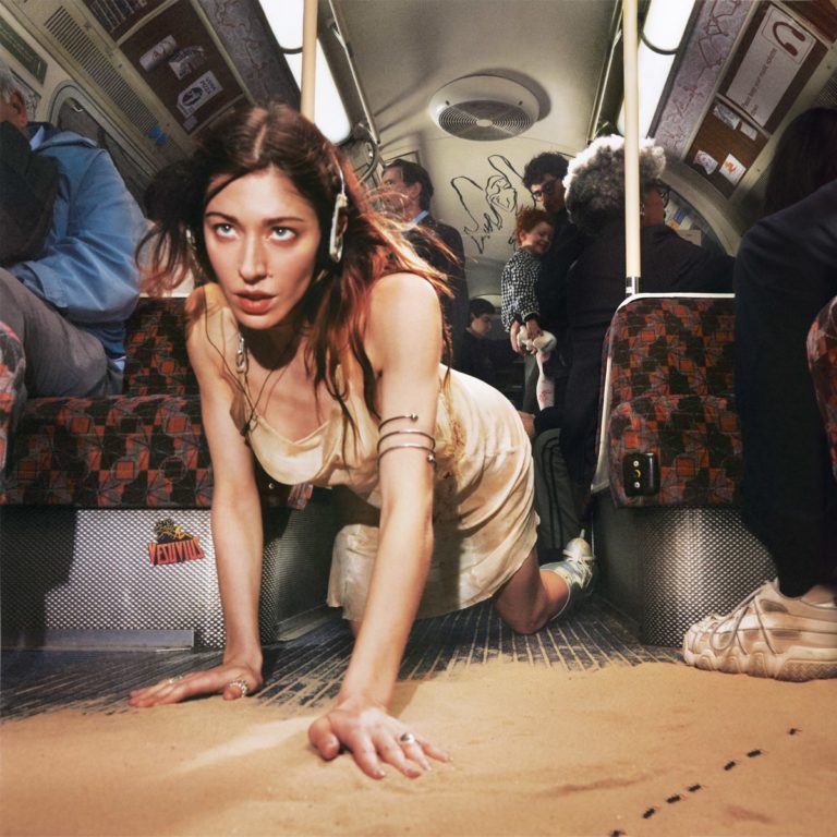 Cover des Albums „Desire, I Want To Turn Into You“ von Caroline Polachek: Die Künstlerin kriecht durch Sand in einer U-Bahn, mit Kopfhörern im futuristischen Look.