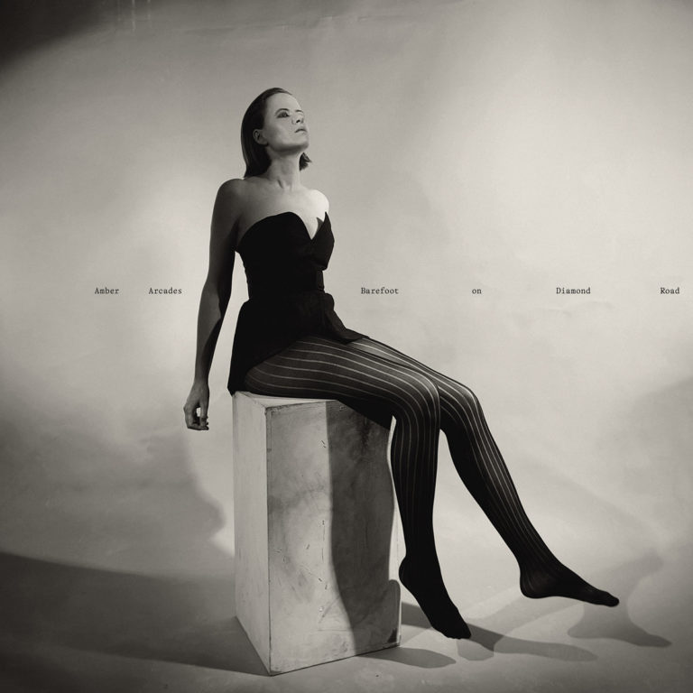 Amber Arcades sitzt in einem stilisierten Fotostudio auf einem weißen Block, trägt ein schwarzes Bustier und gestreifte Strumpfhosen – Cover des Albums „Barefoot on Diamond Road“ in monochromer Ästhetik.