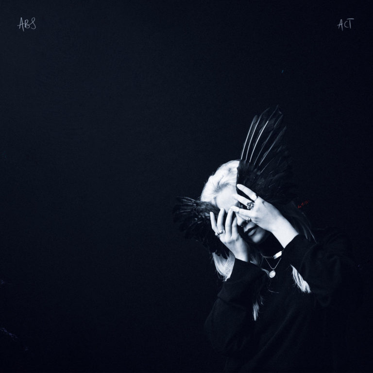 Albumcover von Anna B Savage – A Common Turn: Dunkelblaues, minimalistisches Foto einer Frau mit hellen Haaren, die ihr Gesicht teilweise mit gespreizten schwarzen Vogelflügeln verdeckt.