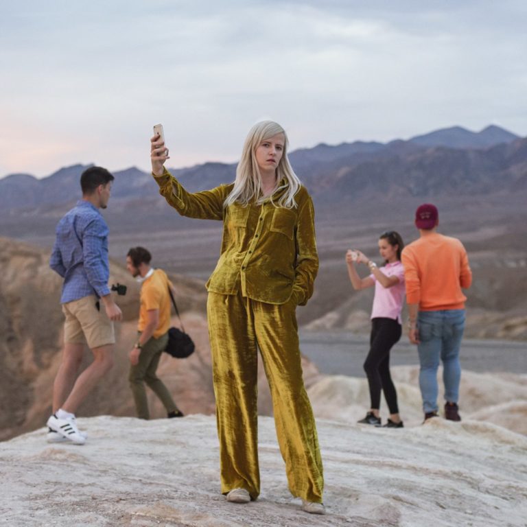 Person in goldener Kleidung macht ein Selfie in einer felsigen Landschaft, umgeben von anderen Menschen.