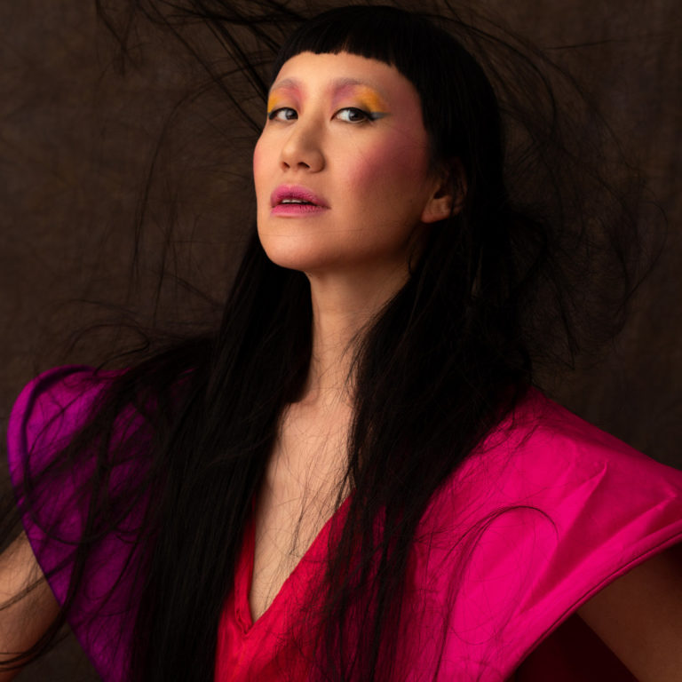 Porträt von Seaming To mit leuchtendem Make-up und avantgardistischem Styling, aufgenommen vor dunklem Hintergrund – Coverbild zum Album „Dust Gatherers“.