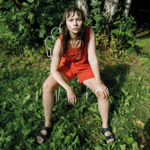 Albumcover von „Gardening“ von Siv Jakobsen mit der Künstlerin in rotem Overall, sitzend auf einem Stuhl im Grünen – introspektiv, roh, naturverbunden.