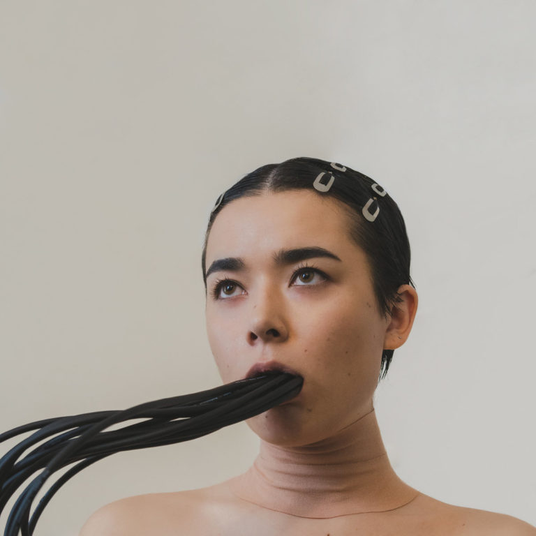 Futuristisches Porträt von Miss Grit mit aus dem Mund ragenden schwarzen Kabeln und minimalistischer Ästhetik – Symbolbild für Cyborg-Identität.