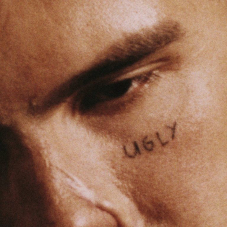 Nahaufnahme eines Gesichts mit dem handschriftlichen Wort „UGLY“ auf der Wange – Cover des Albums „UGLY“ von slowthai, roh und intim.