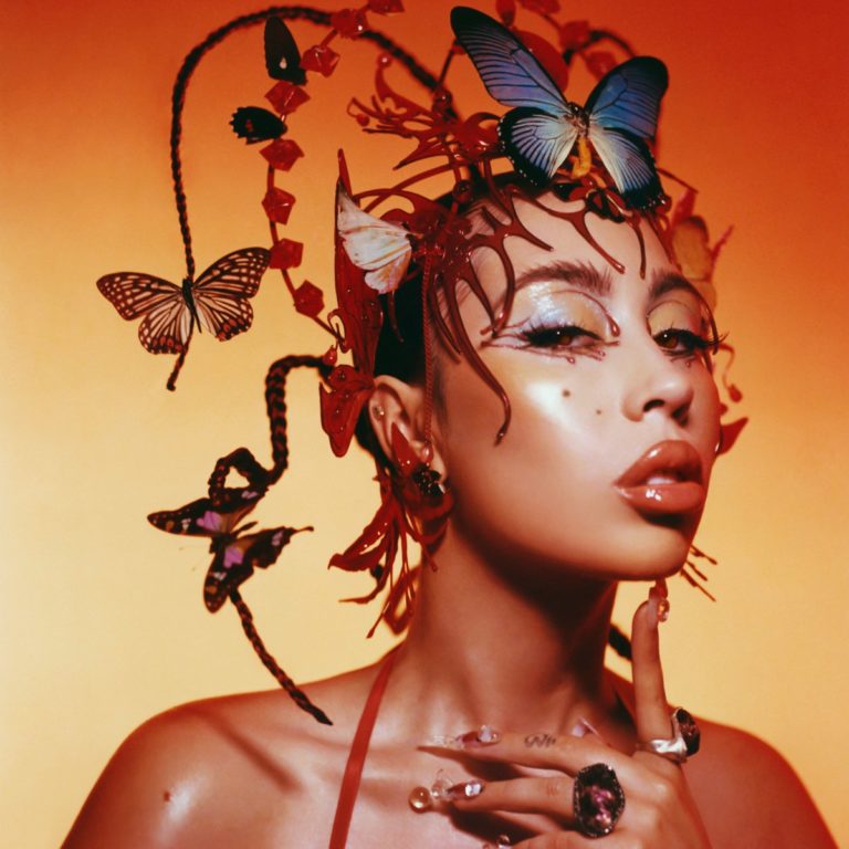 Kali Uchis mit kunstvollem Schmetterlings-Kopfschmuck vor rötlichem Hintergrund – Symbol für das sinnlich-surrealistische Album „Red Moon in Venus“.