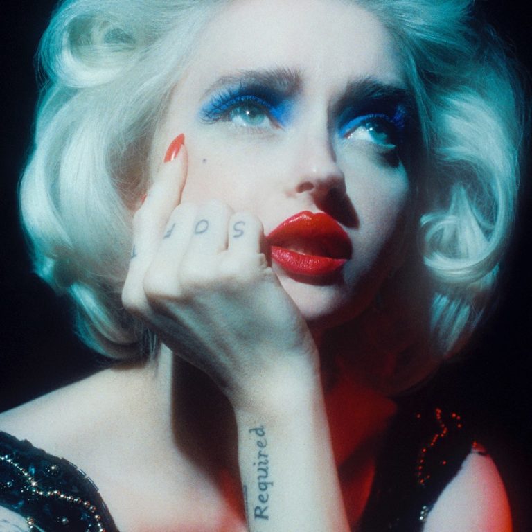 Nahaufnahme einer Frau mit platinblondem Haar, intensiver blauer Lidschatten und rotem Lippenstift; nostalgisch ins Licht blickend, Tattoos an Hand und Arm sichtbar