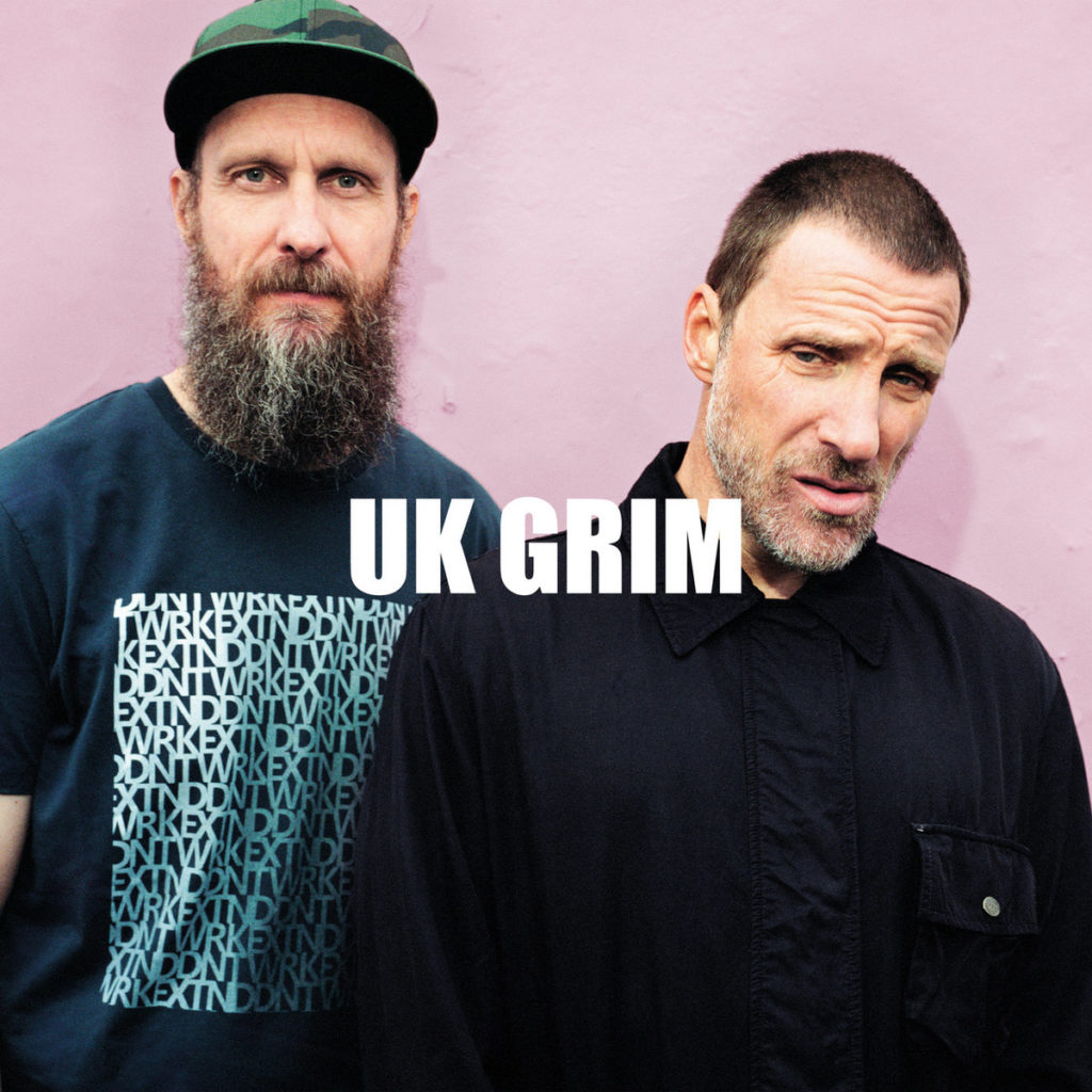 Cover des Albums „UK GRIM“ von Sleaford Mods mit den beiden Bandmitgliedern vor rosa Wand, schlichte Pose, direkter Blick, weißer Albumtitel mittig platziert.