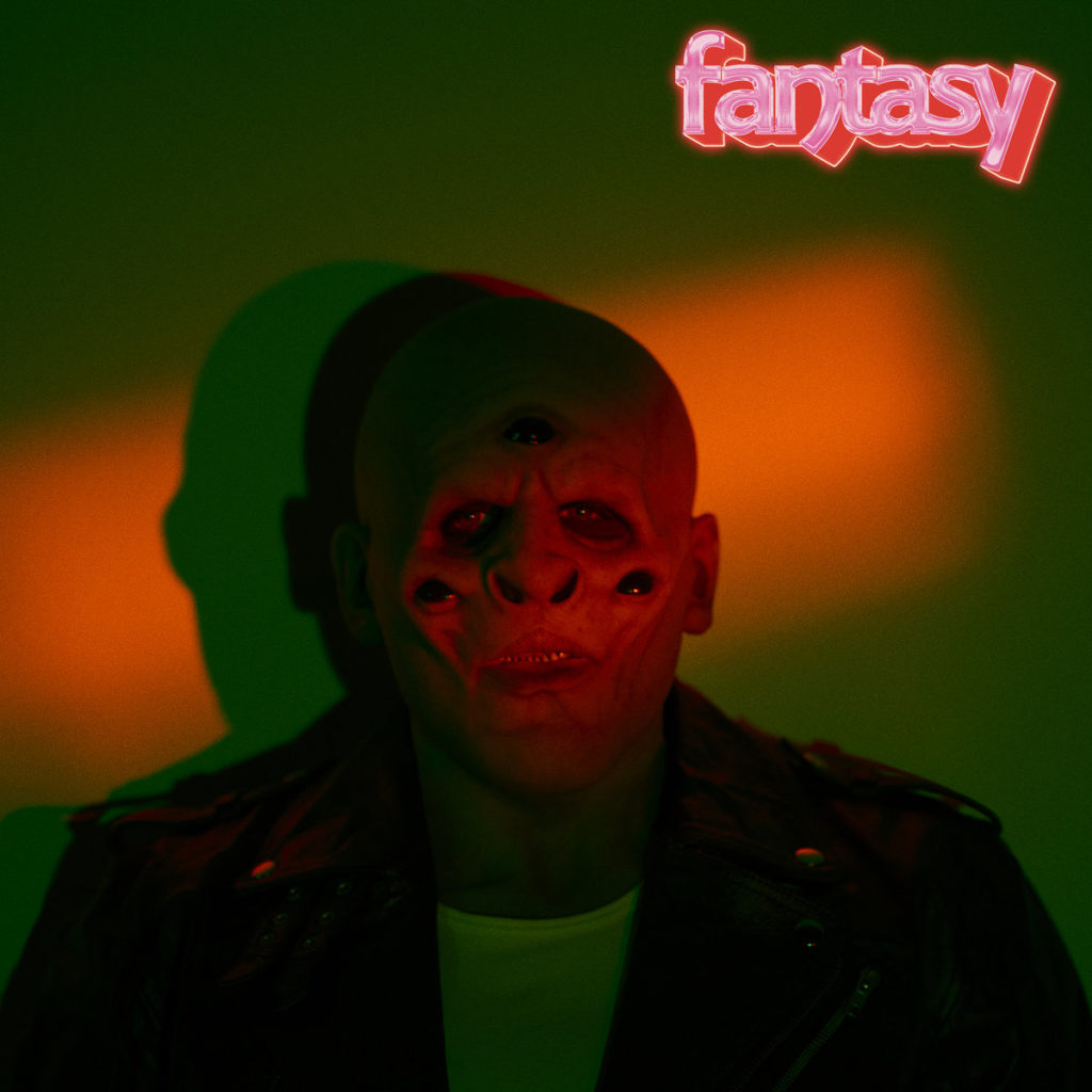Albumcover von „Fantasy“ von M83: Eine surreale Figur mit mehreren Augen und Mündern im Gesicht steht im grünen und roten Licht, darüber leuchtet der Schriftzug „fantasy“.
