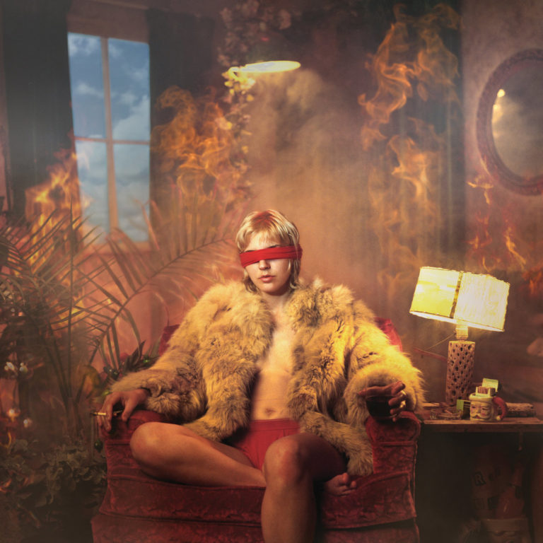 Albumcover von „The Art of Forgetting“ von Caroline Rose: Eine Person mit Felljacke und Augenbinde sitzt in einem brennenden, surrealen Wohnzimmer auf einem Sessel.