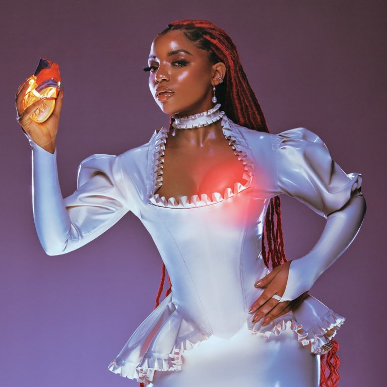 Albumcover von Chlöe – In Pieces: Die Künstlerin in futuristischem, silbernem Kleid mit leuchtendem Herzmotiv, hält ein anatomisches Herz vor violettem Hintergrund.