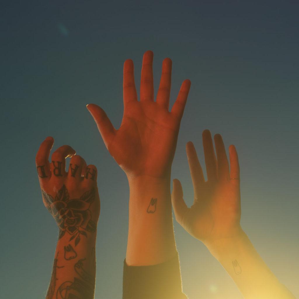 Albumcover von Boygenius – the record: Drei erhobene Hände vor blauem Himmel im Sonnenlicht, mit Zahn-Tattoos und Schriftzug „HARD“ auf Fingern, symbolisch und minimalistisch.