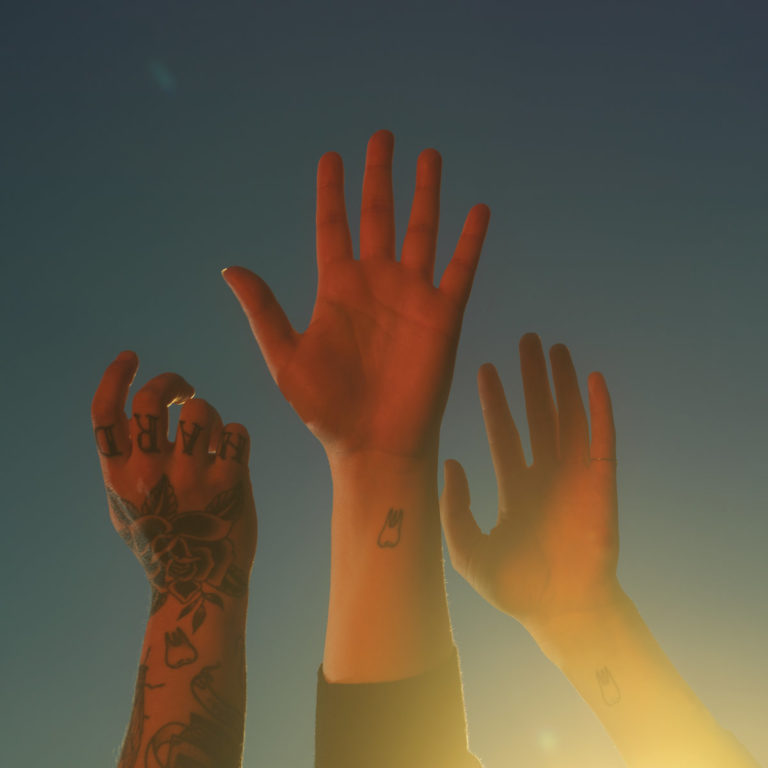 Albumcover von Boygenius – the record: Drei erhobene Hände vor blauem Himmel im Sonnenlicht, mit Zahn-Tattoos und Schriftzug „HARD“ auf Fingern, symbolisch und minimalistisch.