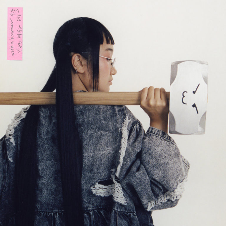 Albumcover von "With A Hammer" von Yaeji zeigt die Künstlerin seitlich mit einem stilisierten Hammer über der Schulter, vor hellem Hintergrund.