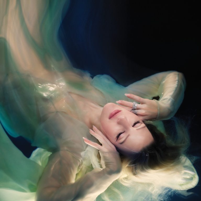 Albumcover von "Higher Than Heaven" von Ellie Goulding mit schwebender Pose unter Wasser, verträumtem Gesichtsausdruck und fließendem Kleid in Pastellfarben.
