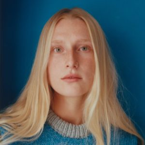 Portraitfoto von Billie Marten vor blauem Hintergrund, mit natürlichem Licht, blondem Haar und gestricktem Pullover in Blau und Grau.