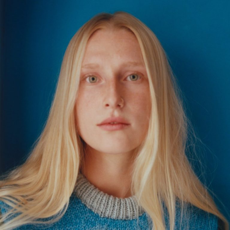 Portraitfoto von Billie Marten vor blauem Hintergrund, mit natürlichem Licht, blondem Haar und gestricktem Pullover in Blau und Grau.
