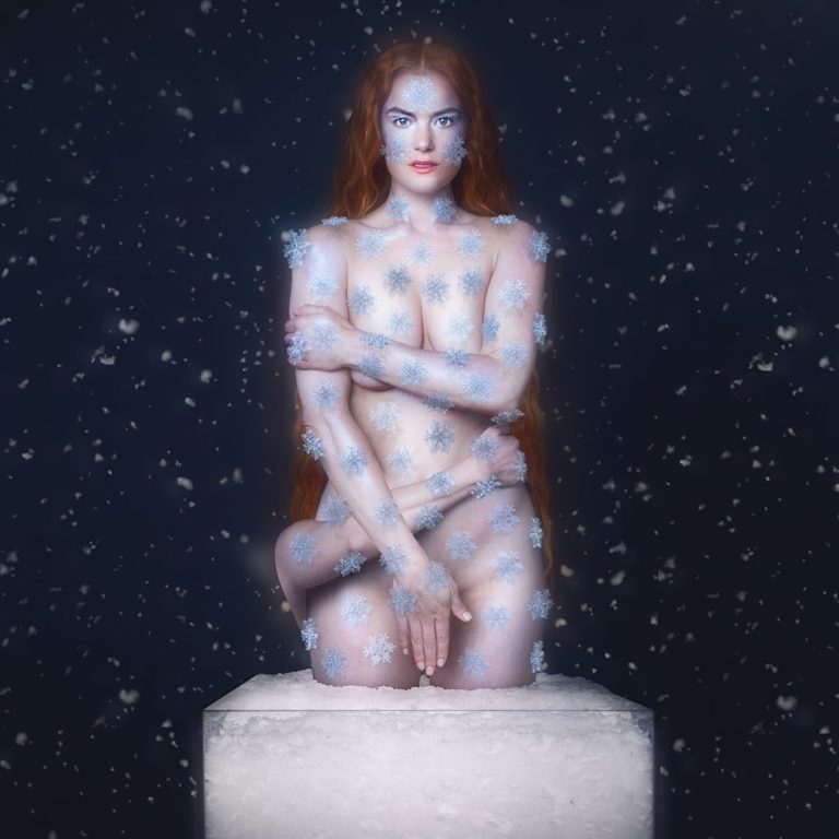 Julia Stone kniet nackt auf einem Eisblock, bedeckt mit funkelnden Schneeflocken, während dichter Schneefall vor dunklem Hintergrund ihre rotblonden Haare umrahmt – eine surreale, frostige Inszenierung zwischen Zerbrechlichkeit und Glanz.