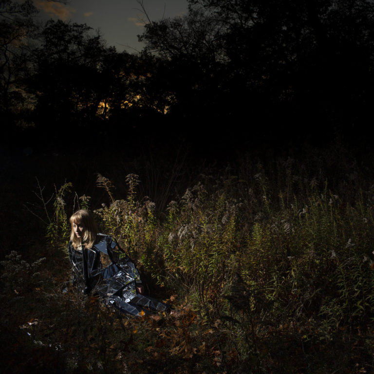 Albumcover zu Ignorance von The Weather Station: Eine Frau sitzt bei Dämmerung in einer verwilderten Lichtung, eingehüllt in eine reflektierende silberne Folie; düstere Natur, melancholische Atmosphäre.