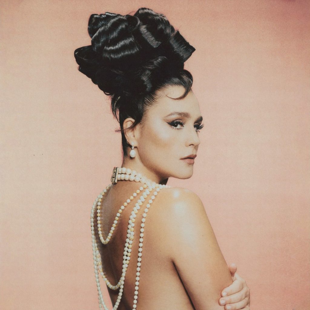 Jessie Ware mit kunstvoll aufgestecktem Haar, Perlenketten auf dem nackten Rücken, vor rosafarbenem Hintergrund – retro-glamourös inszeniert.