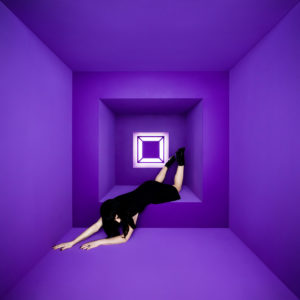 Eine Frau in schwarzem Kleid liegt surreal an der Wand eines vollständig lilafarbenen Würfelraums; Licht strömt aus einem geometrischen Fenster im Hintergrund.