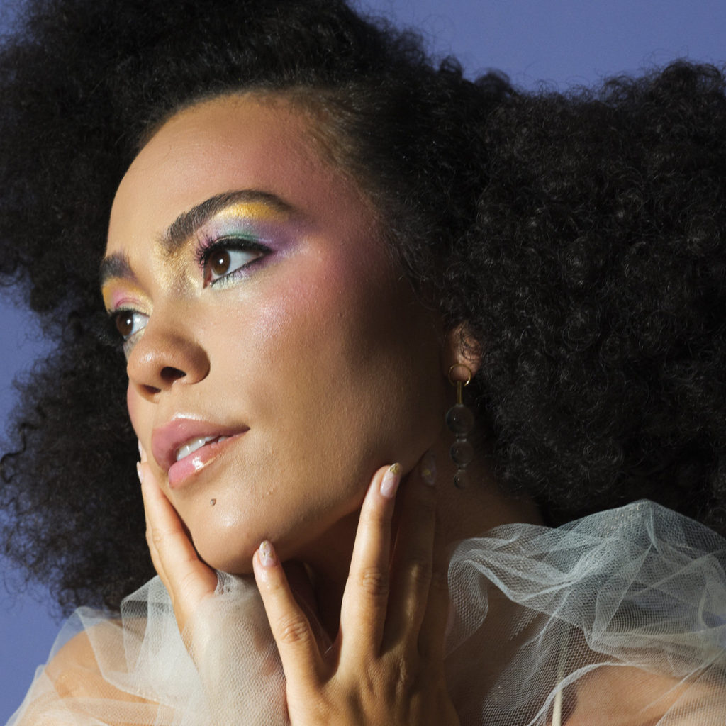 Nahaufnahme von Madison McFerrin mit farbigem Make-up, durchscheinendem Tüll und verträumtem Blick vor lilafarbenem Hintergrund.