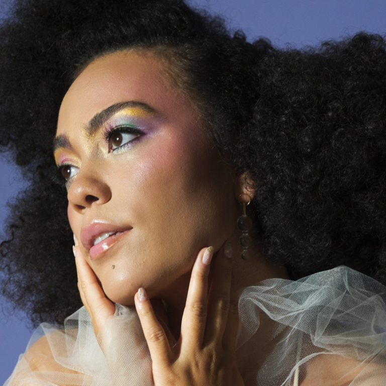 Nahaufnahme von Madison McFerrin mit farbigem Make-up, durchscheinendem Tüll und verträumtem Blick vor lilafarbenem Hintergrund.
