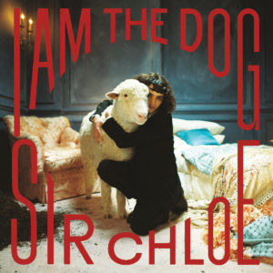 Dana Foote von Sir Chloe umarmt ein Schaf in einem opulenten, leicht chaotischen Zimmer – auf dem Cover von „I Am The Dog“ in roten Lettern.