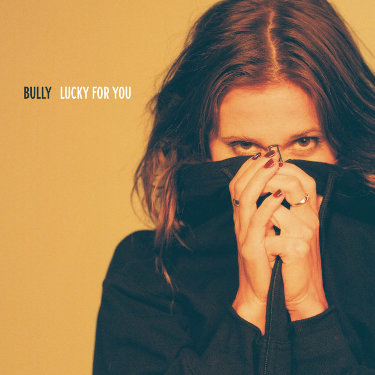 Cover des Albums Lucky For You von Bully: Frau verdeckt teilweise ihr Gesicht mit schwarzem Hoodie, gelblicher Hintergrund, intensiver Blick.