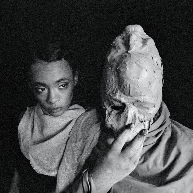Cover von Formal Growth in the Desert von Protomartyr: Schwarzweiß-Foto mit junger Frau und Figur mit grober Maske – düster, mystisch, nachdenklich.
