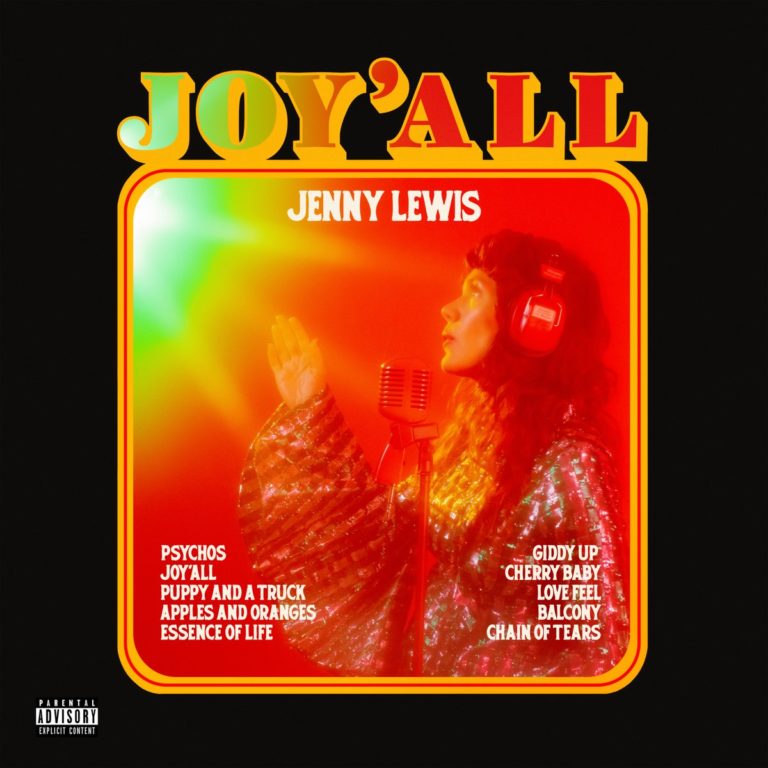 Cover des Albums „Joy’All“ von Jenny Lewis – die Künstlerin singt ins Mikrofon, trägt glitzerndes Outfit und rote Kopfhörer, in warmem, retrofarbenem Studiolicht.