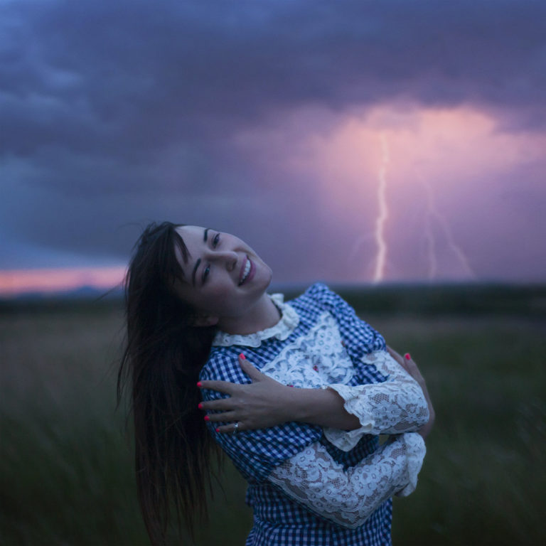 Albumcover von „Time Ain’t Accidental“ – Jess Williamson steht in einem Feld, umarmt sich selbst im viktorianisch inspirierten Kleid, hinter ihr ein dramatischer Himmel mit Blitz.